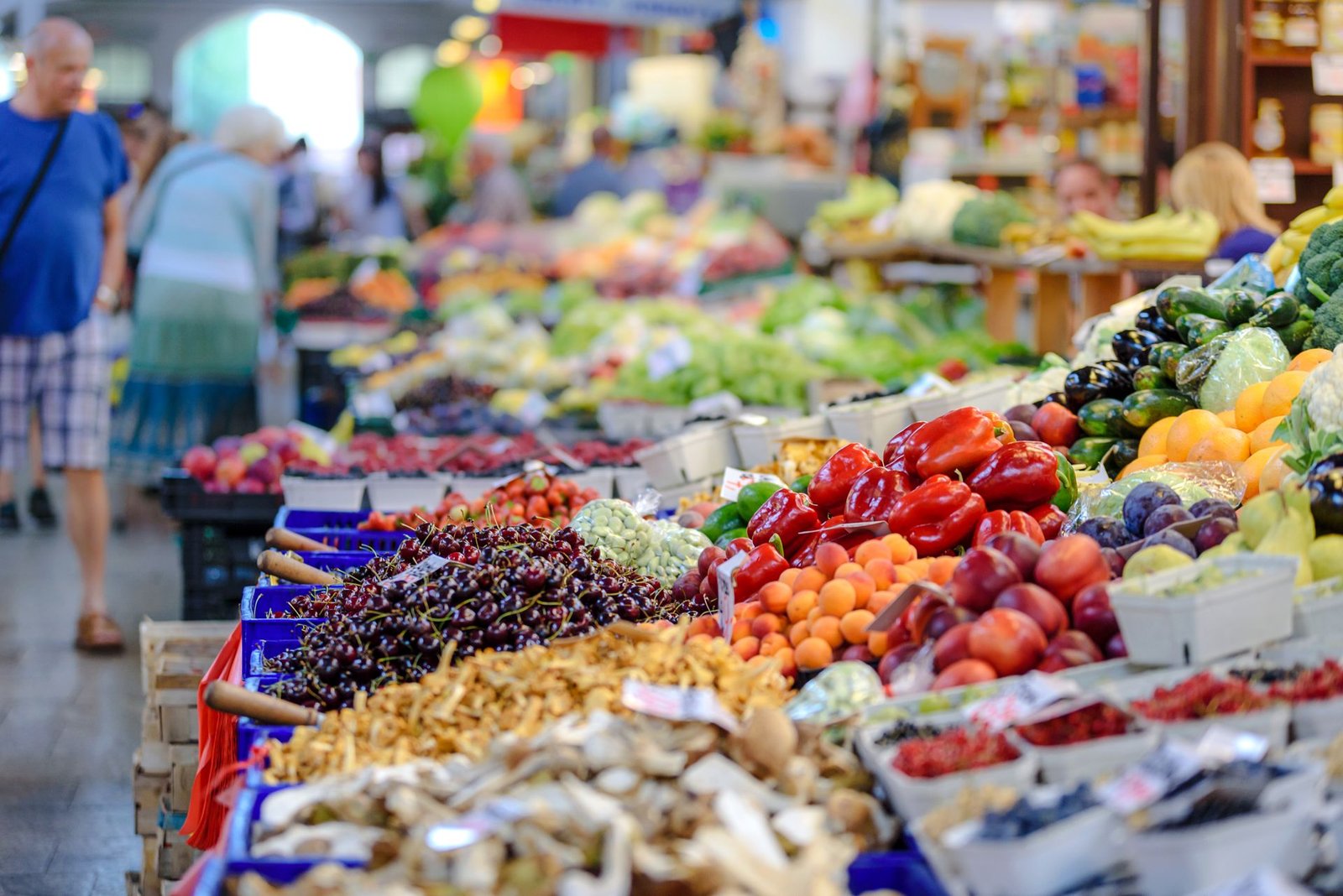 Bancas de feira com frutas, legumes e cogumelos expostos para venda, com clientes ao fundo desfocados