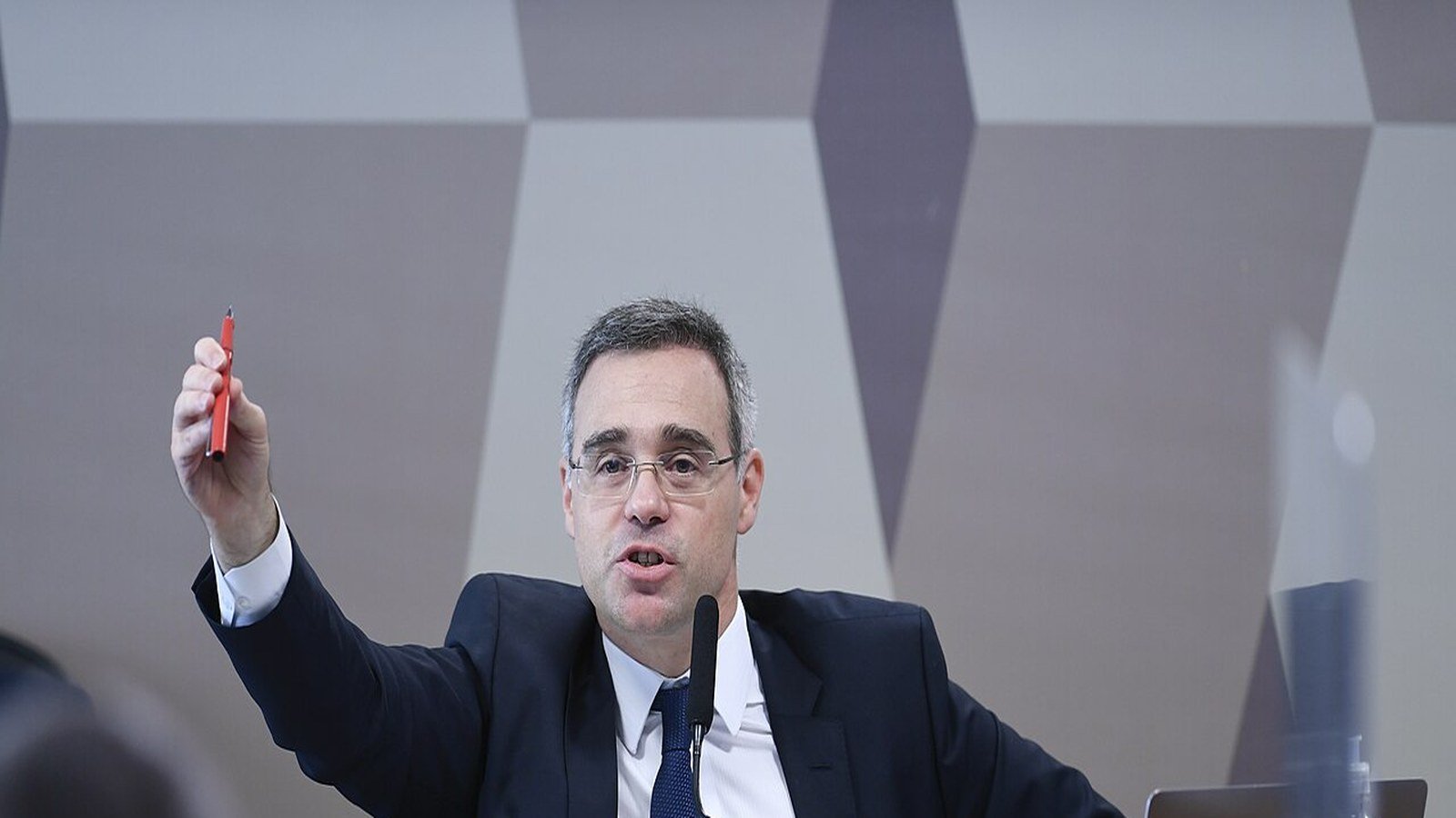 O ministro André Mendonça fala ao microfone, de terno escuro, com uma caneta na mão erguida