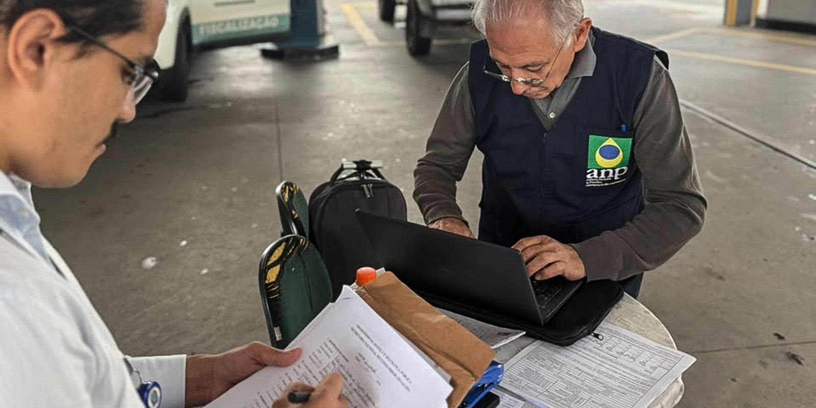 Dois fiscais conferem documentos em um posto de combustíveis; um deles usa colete com o logotipo da ANP