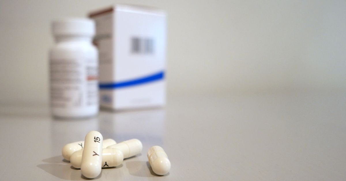 Cápsulas brancas sobre superfície clara, com frasco e caixa de medicamento desfocados ao fundo