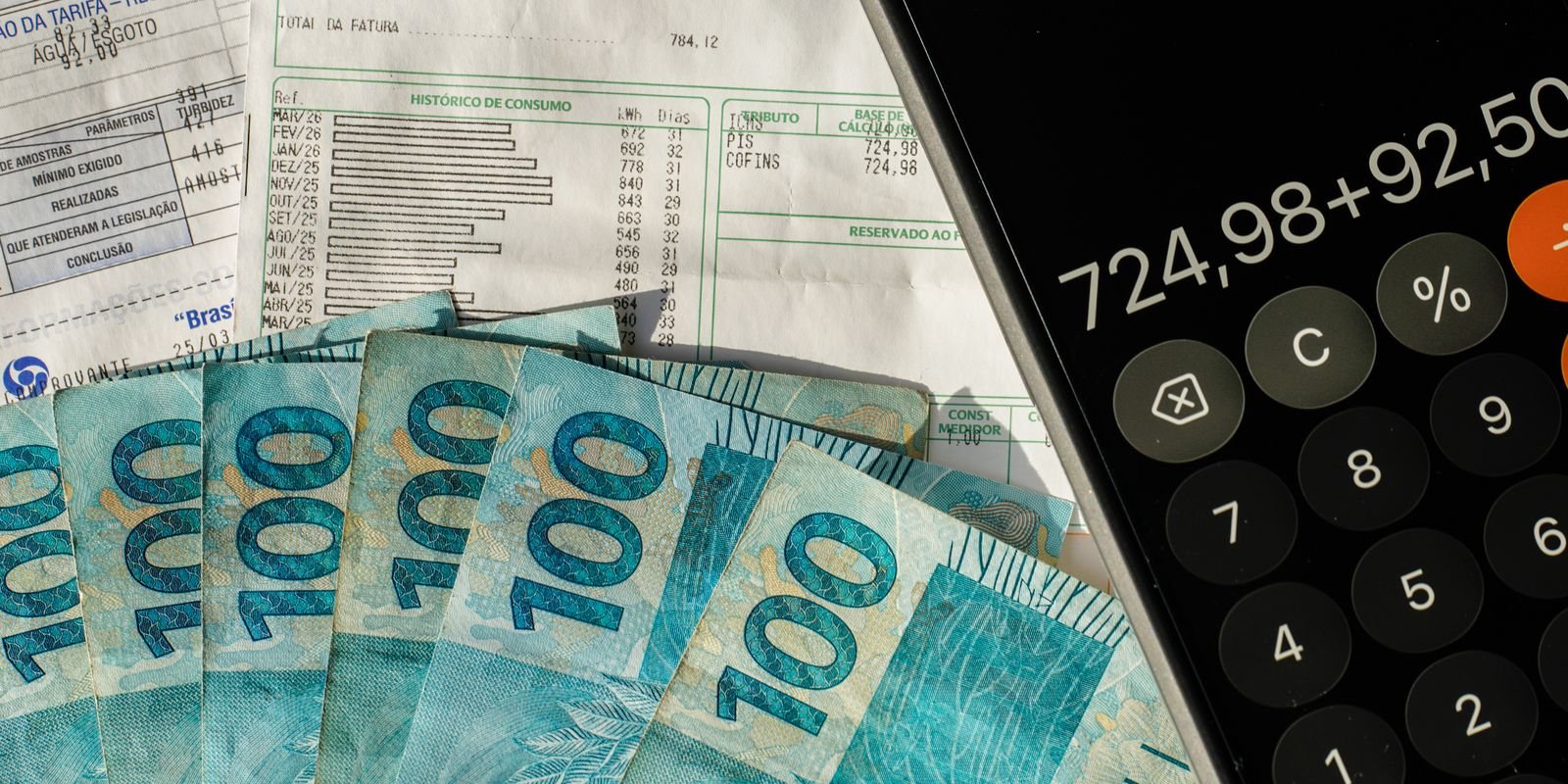 Notas de R$ 100 sobre contas de luz e água, ao lado de uma calculadora de celular