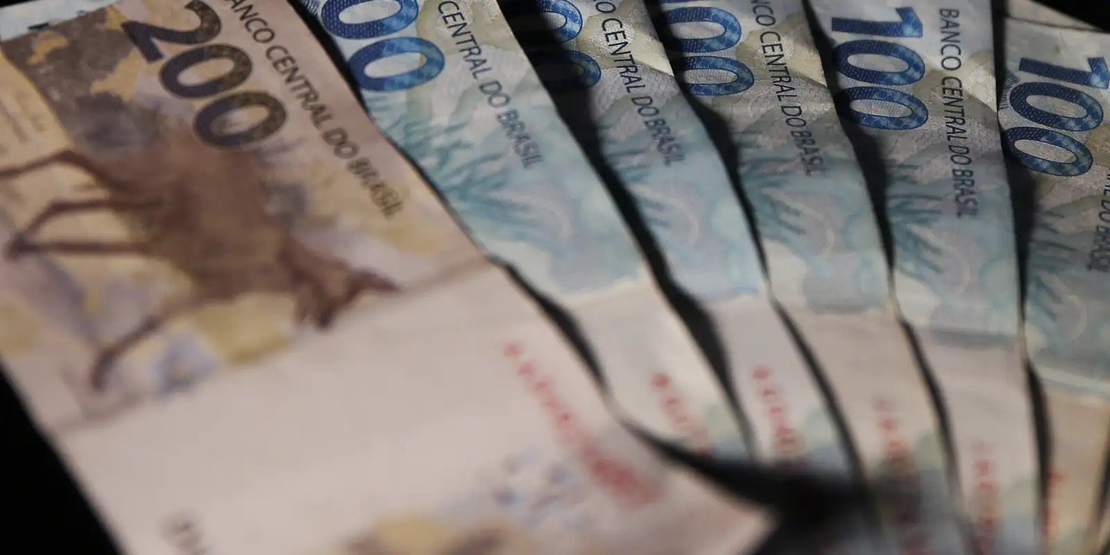 Cédulas de 100 e 200 reais enfileiradas em close, fotografadas em ângulo