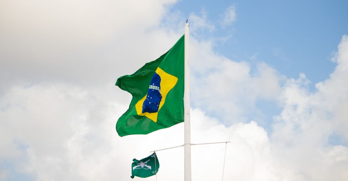 Bandeira do Brasil hasteada em mastro branco contra o ceu com nuvens