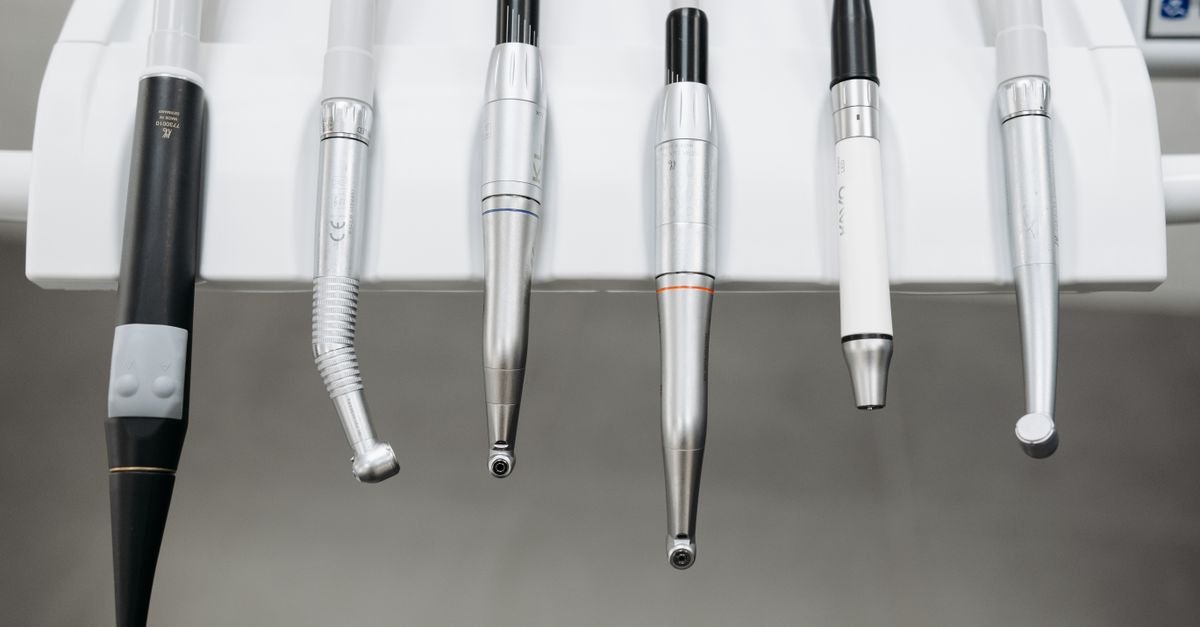 Seis instrumentos odontológicos metálicos pendurados lado a lado no suporte de um equipamento de consultório