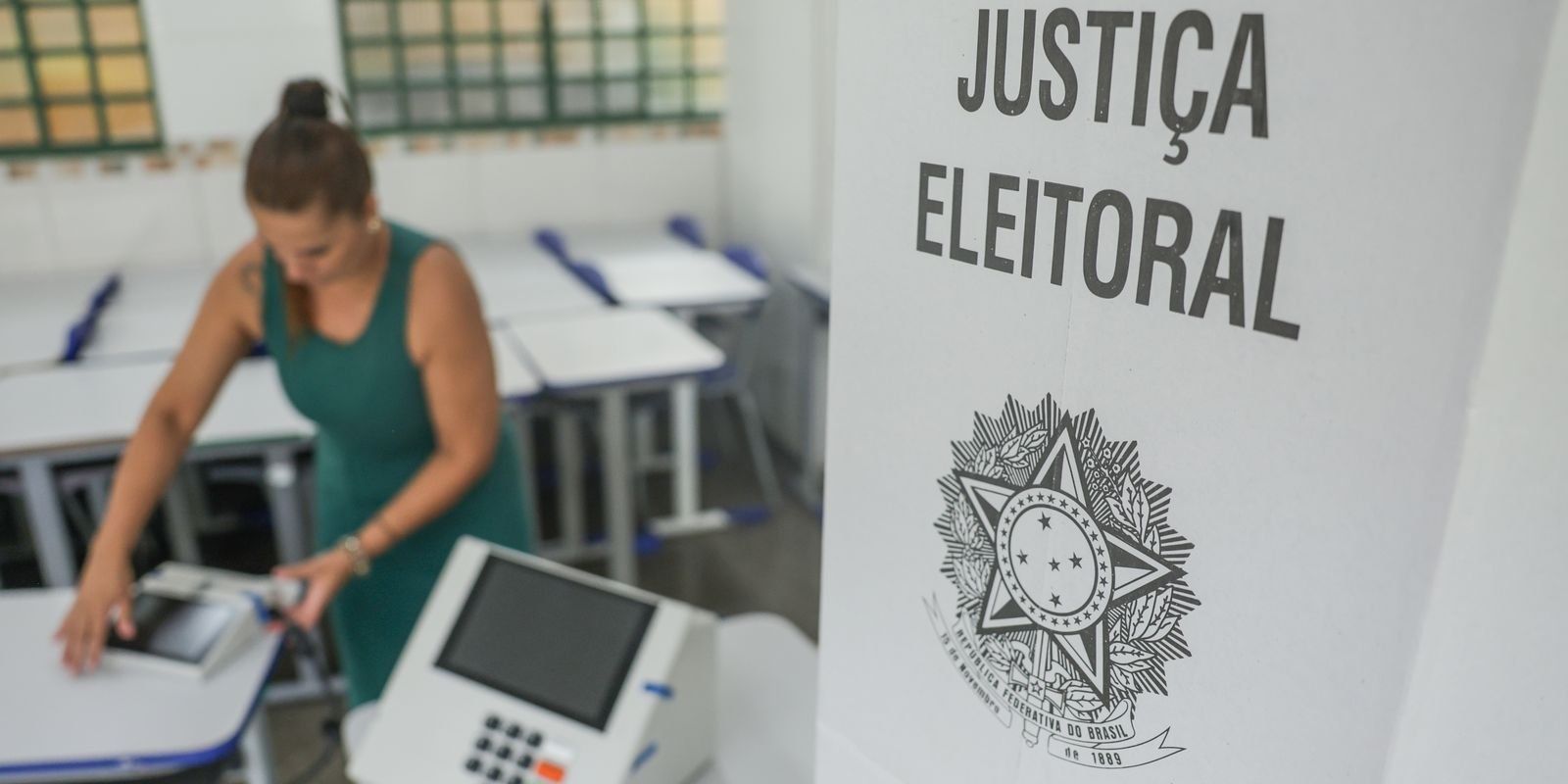 Urna eletrônica sendo preparada em sala de aula ao lado de cartaz da Justiça Eleitoral