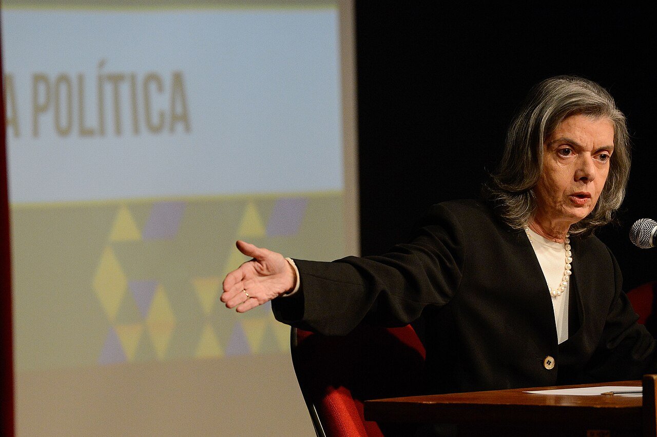 A ministra Carmen Lucia fala ao microfone durante evento, com tela de projecao ao fundo