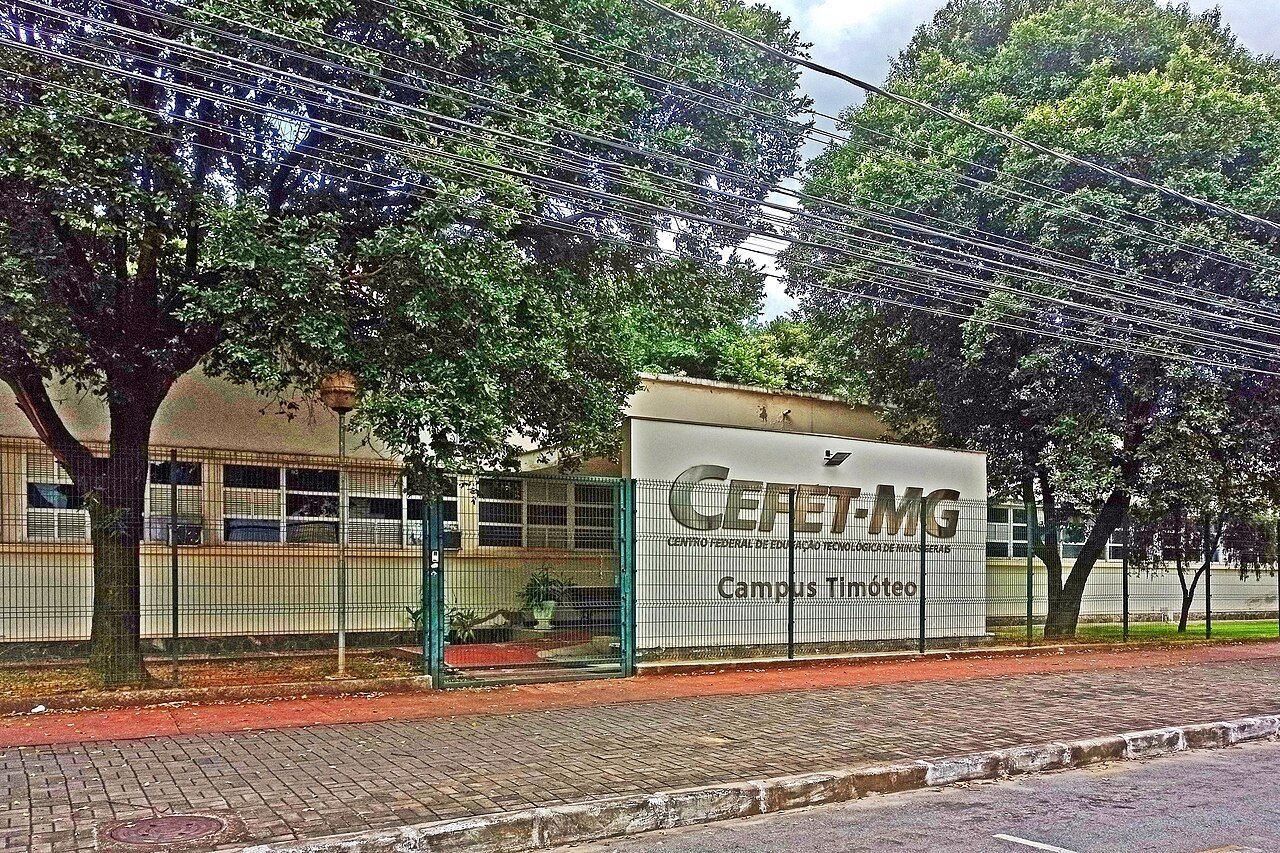Fachada de campus do CEFET-MG com o letreiro da instituicao visivel atras da grade, em rua arborizada