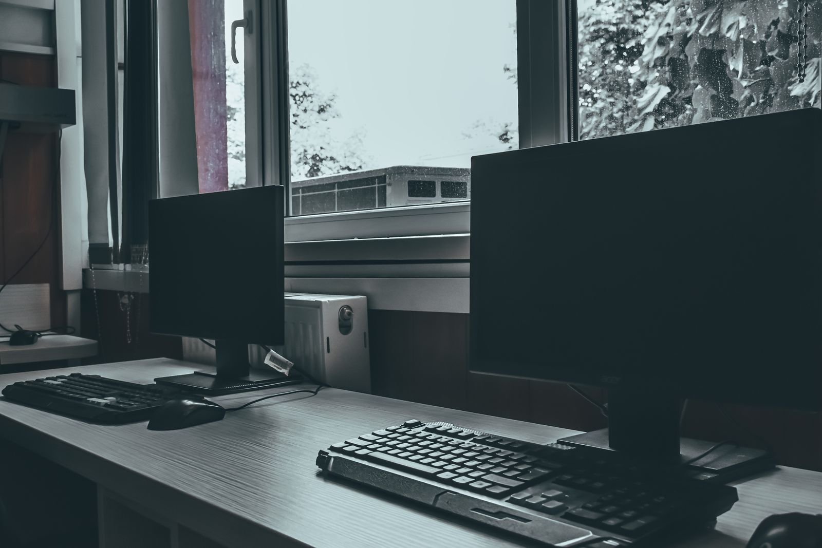 Mesa com dois monitores desligados e teclado diante de uma janela