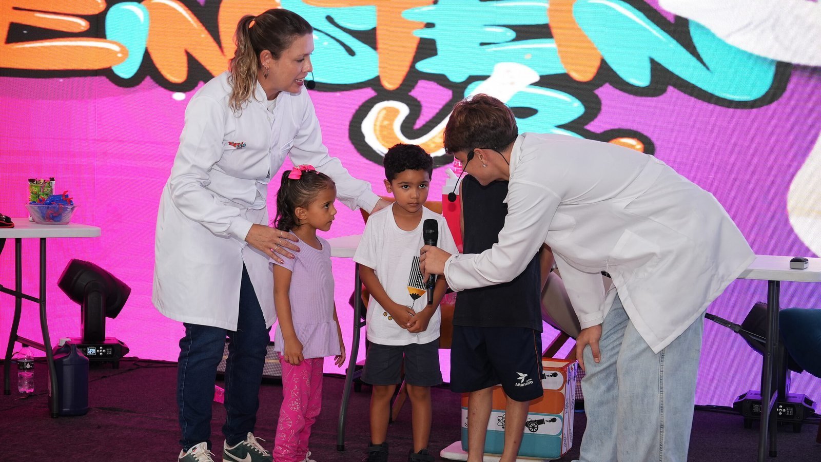 Monitores de jaleco branco conduzem três crianças em experimento no palco do Einstein Jr.
