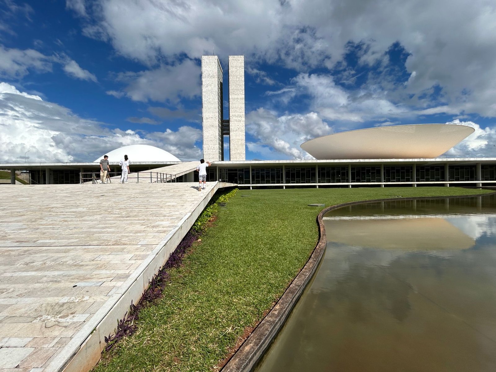 Torres e cúpulas do Congresso Nacional em Brasília sob céu azul, com o espelho d'água em primeiro plano