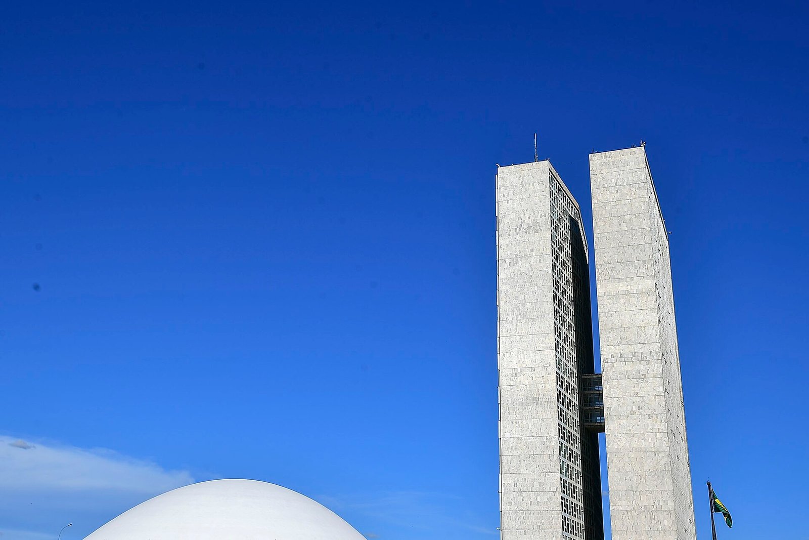 Torres do Congresso Nacional e a cúpula do plenário do Senado vistas de baixo, com céu azul e a bandeira do Brasil