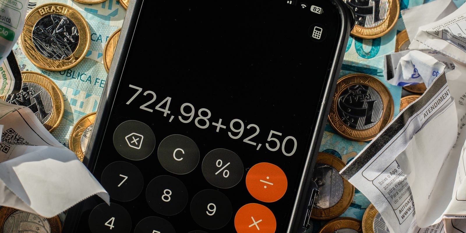 Celular com aplicativo de calculadora aberto sobre moedas e cédulas de real e cupons fiscais amassados