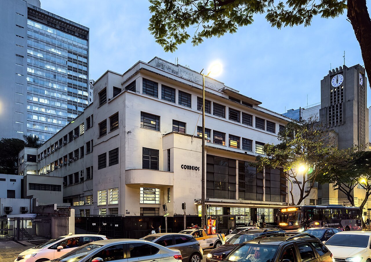 Fachada de agência central dos Correios com o letreiro da empresa, movimento de carros e ônibus na via em frente