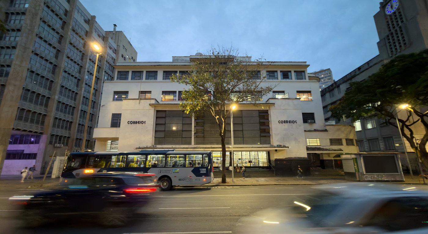 Fachada da Agência Central dos Correios de Belo Horizonte ao anoitecer, com ônibus e carros passando na avenida