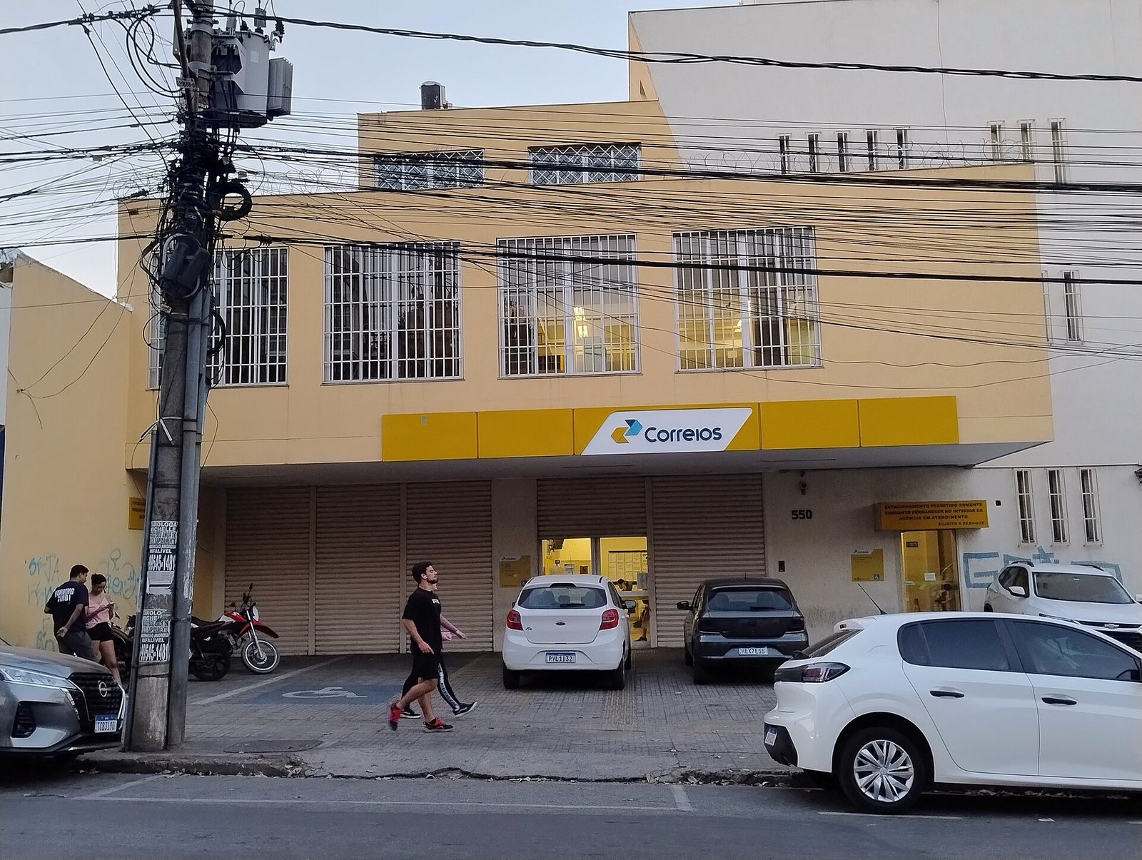 Fachada de uma agência dos Correios com o letreiro amarelo sobre a entrada e carros estacionados na frente