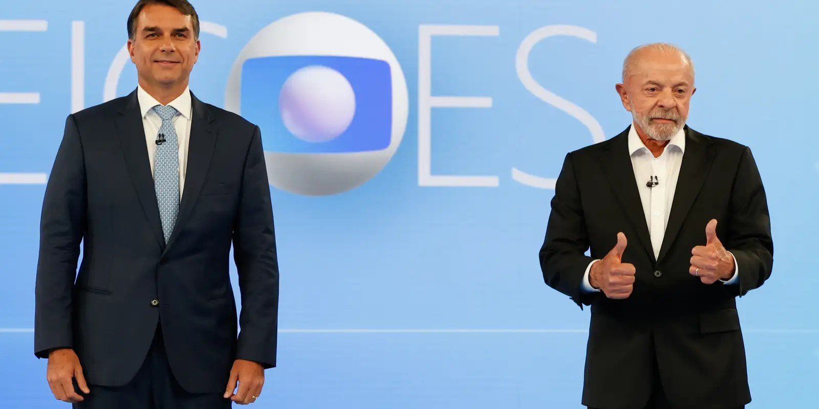 Flávio Bolsonaro e Lula lado a lado no estúdio do debate da TV Globo, diante do cenário azul do programa