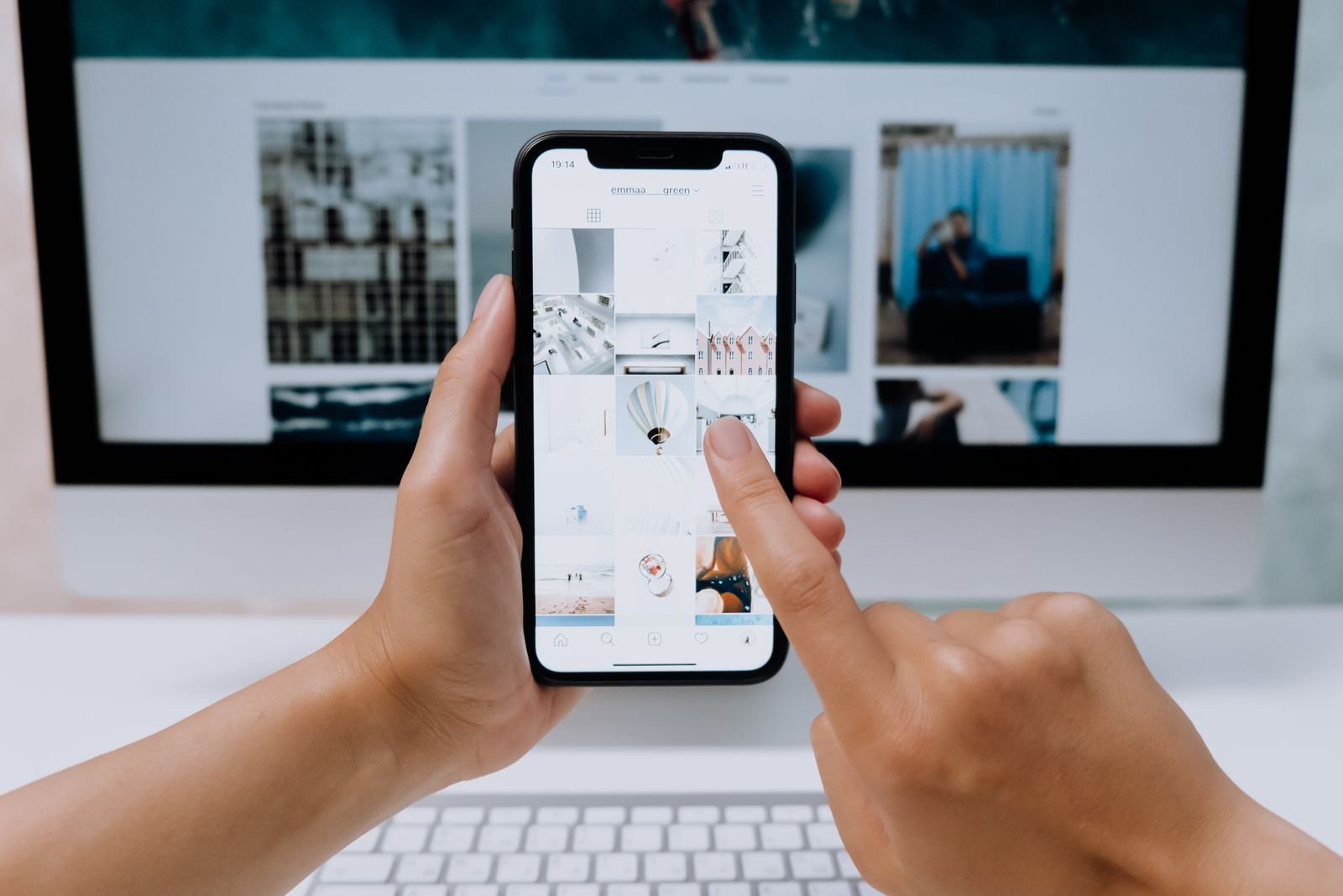 Mãos seguram um celular com um feed de rede social aberto diante de um computador