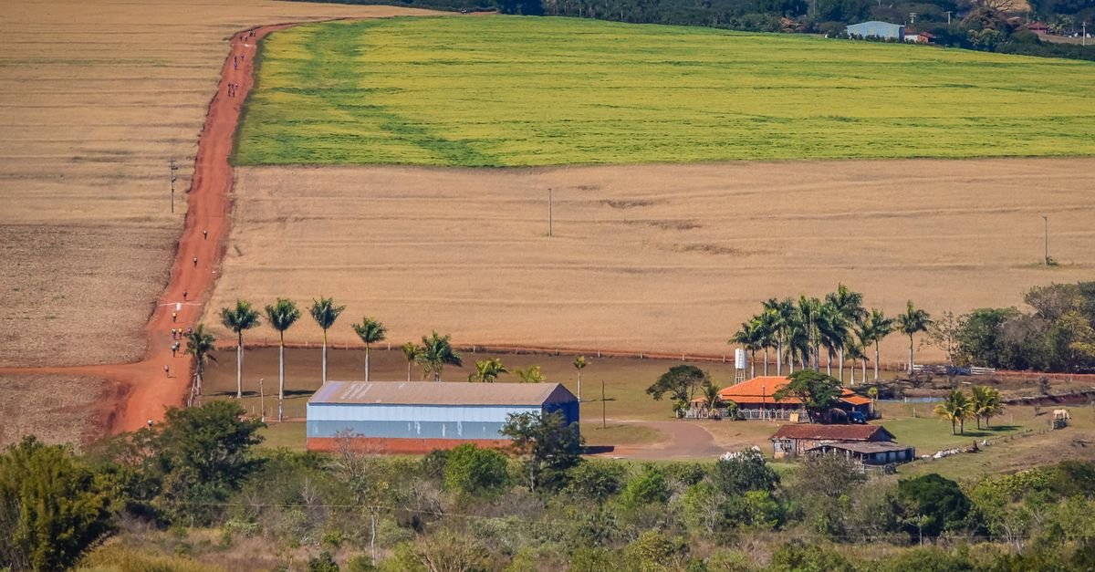 Vista aérea de propriedade rural com lavouras, estrada de terra vermelha, galpão e sede da fazenda