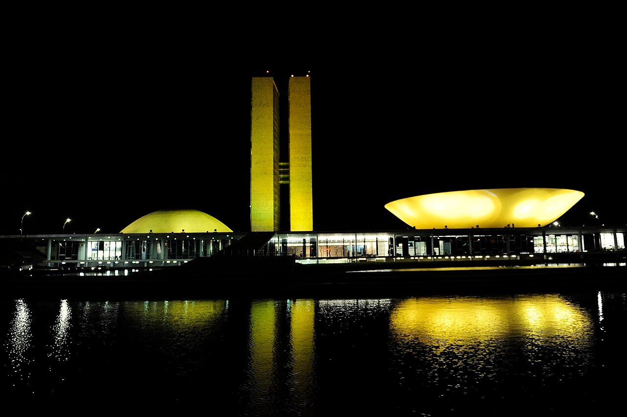 Palácio do Congresso Nacional iluminado à noite, com as cúpulas do Senado e da Câmara refletidas no espelho d'água