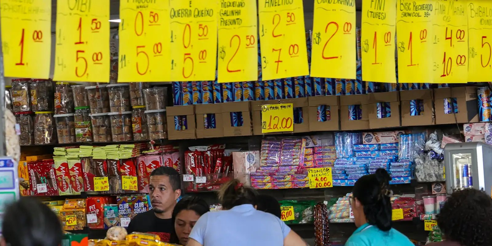 Comerciante atende clientes em pequena loja de bairro com cartazes de preços