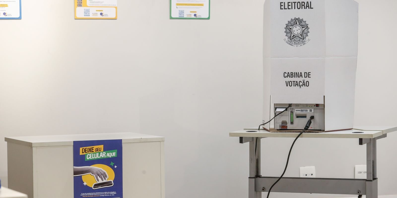 Cabine de votação de papelão da Justiça Eleitoral ao lado de uma urna eletrônica e de um aviso para deixar o celular antes de votar