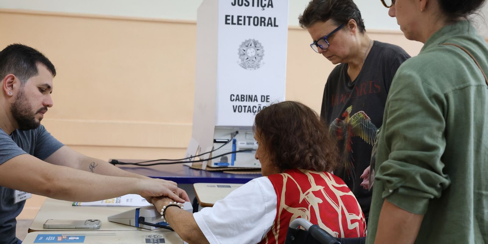 Mesário auxilia eleitora em cadeira de rodas na identificação biométrica diante da cabine de votação, em seção eleitoral no primeiro turno de 2026