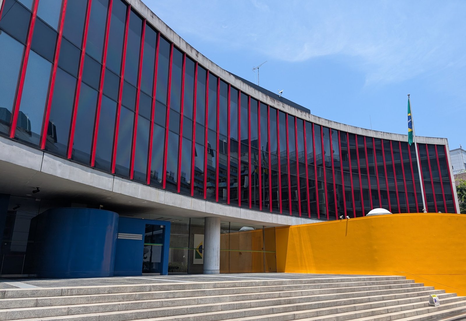 Fachada curva de vidro com montantes vermelhos da Embaixada do Brasil em Tóquio, com a bandeira brasileira no mastro