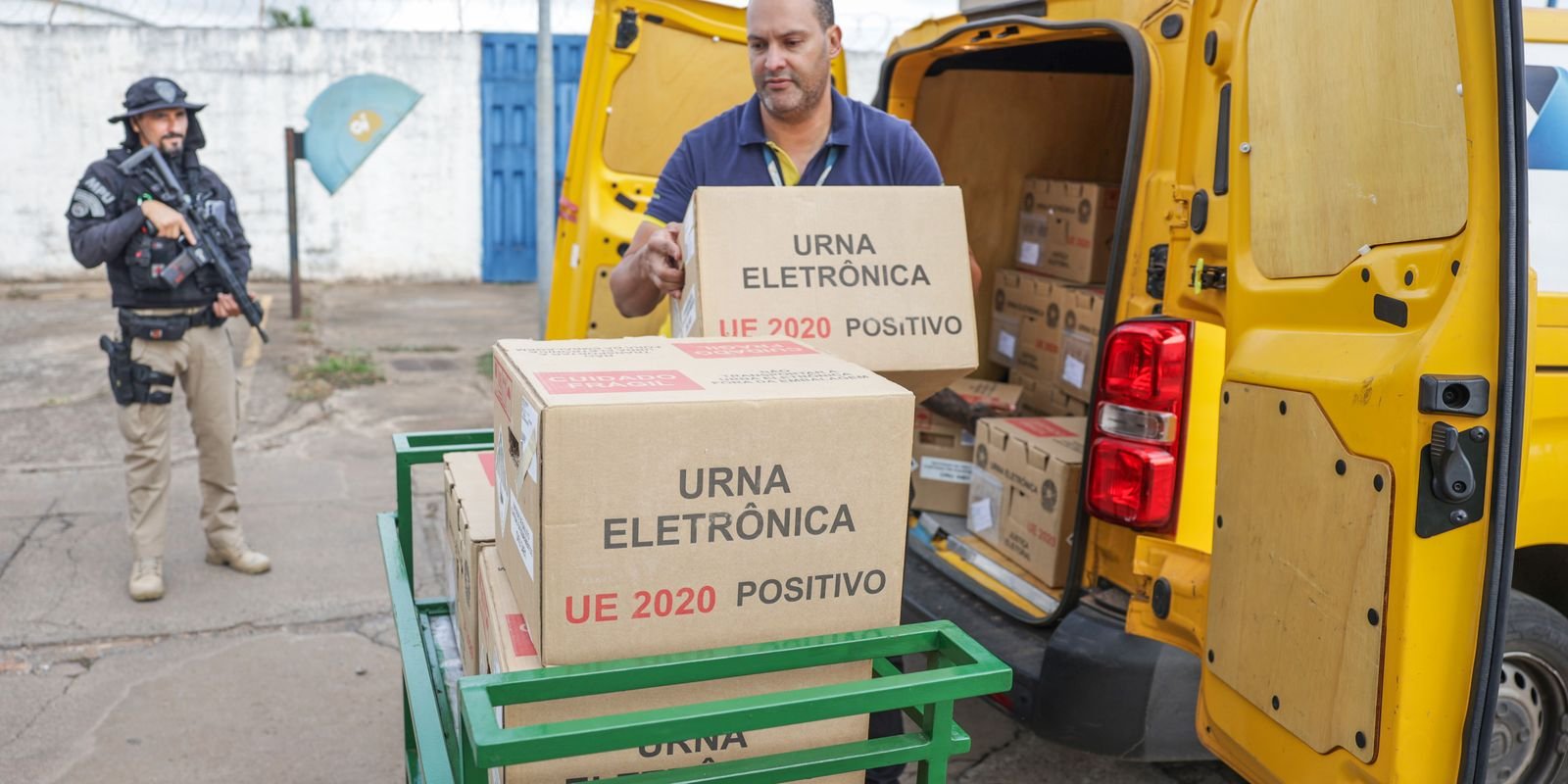 Funcionário da Justiça Eleitoral carrega caixas de urna eletrônica em van enquanto um policial armado acompanha, no Distrito Federal, na distribuição do primeiro turno