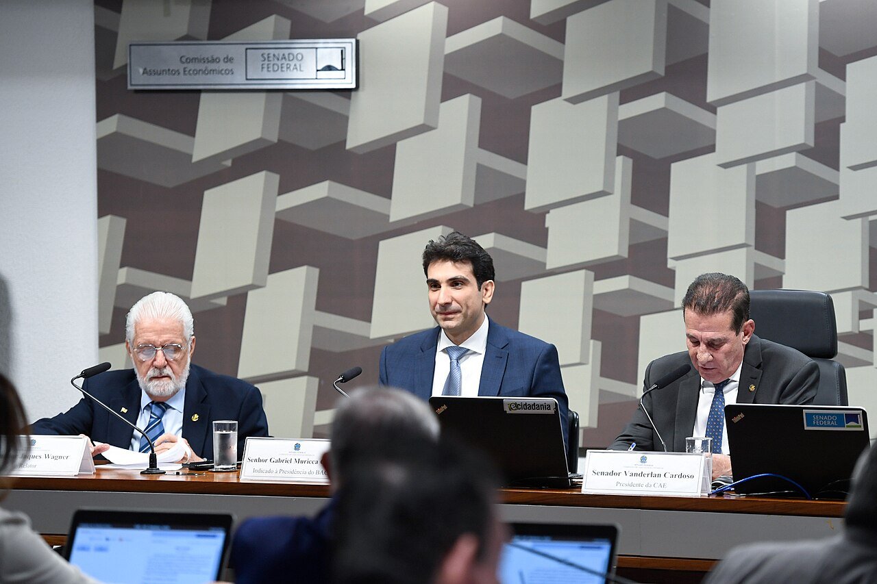 Gabriel Galípolo sentado à mesa da Comissão de Assuntos Econômicos do Senado, entre o relator Jaques Wagner e o presidente da comissão, Vanderlan Cardoso