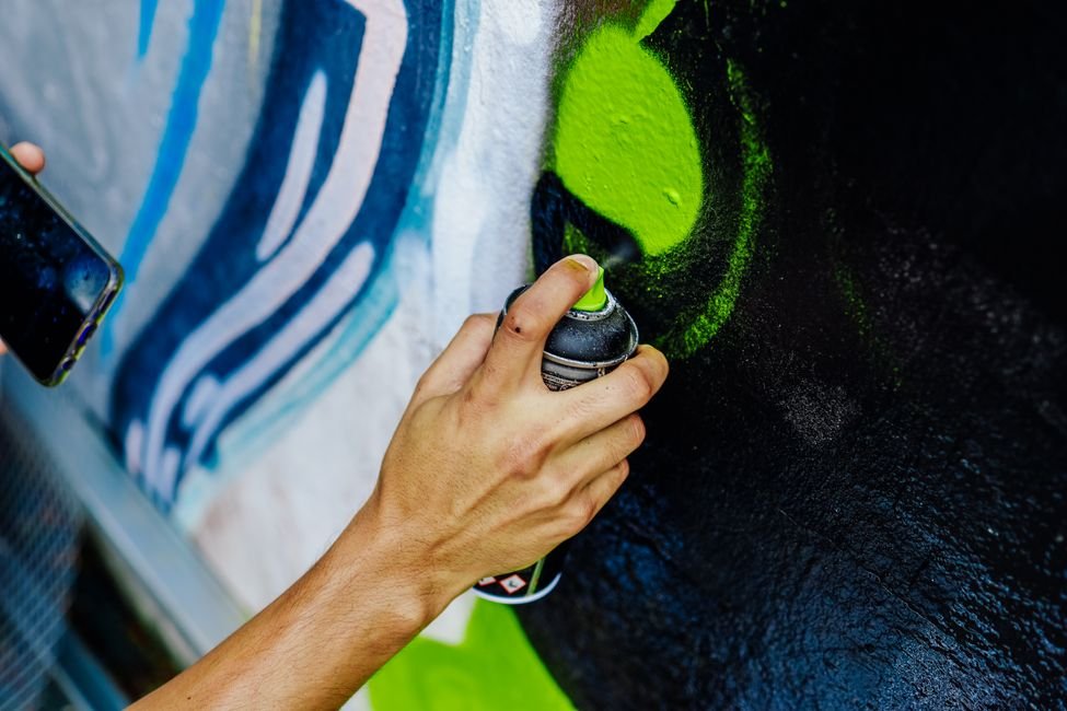 Mao segurando lata de spray aplicando tinta verde sobre um muro grafitado em tons de azul e preto