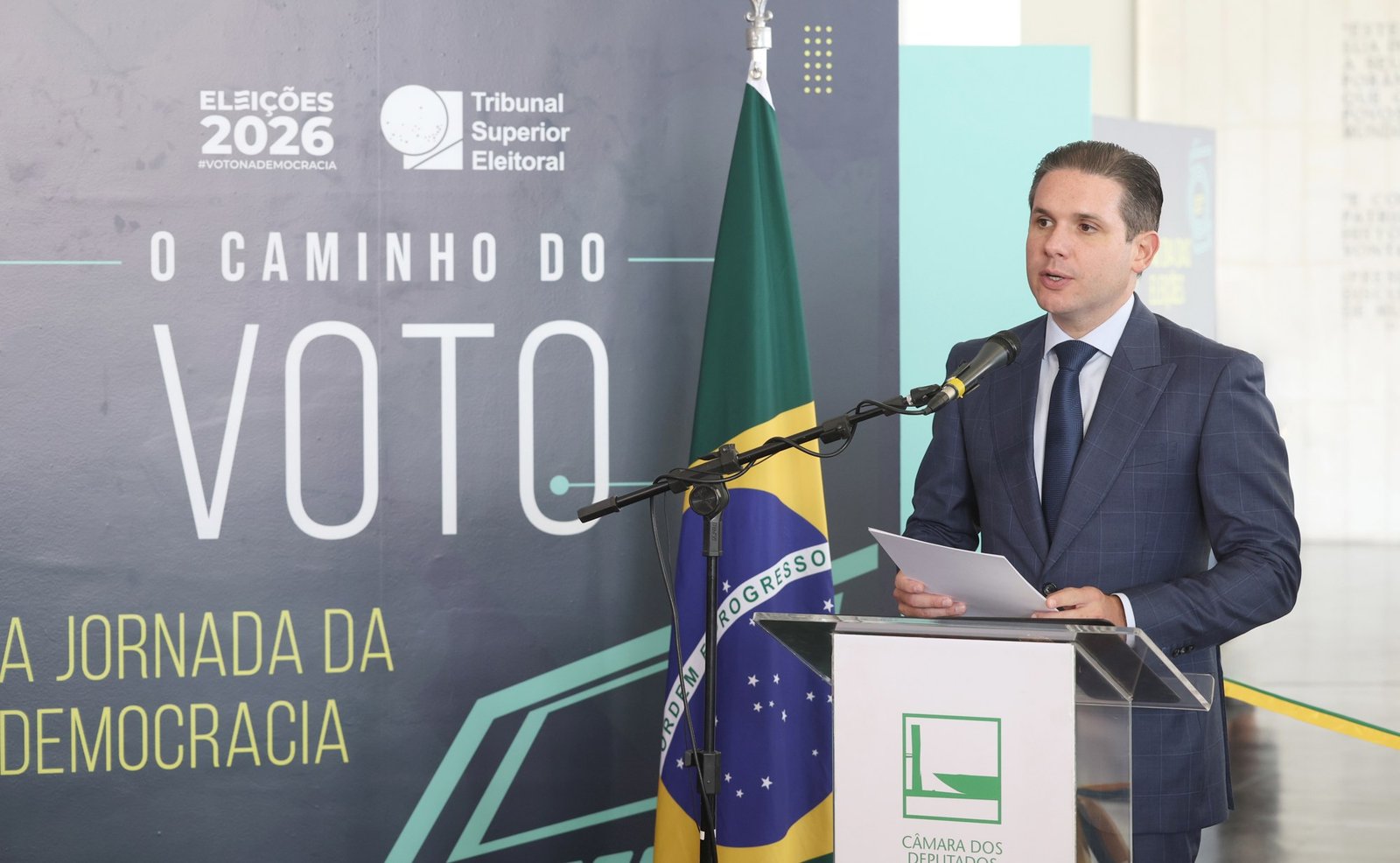 Hugo Motta discursa em púlpito diante de painel da exposição O Caminho do Voto, com a bandeira do Brasil ao lado