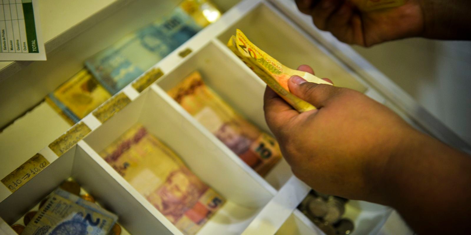 Mãos contam cédulas de real dentro de gaveta de caixa registradora