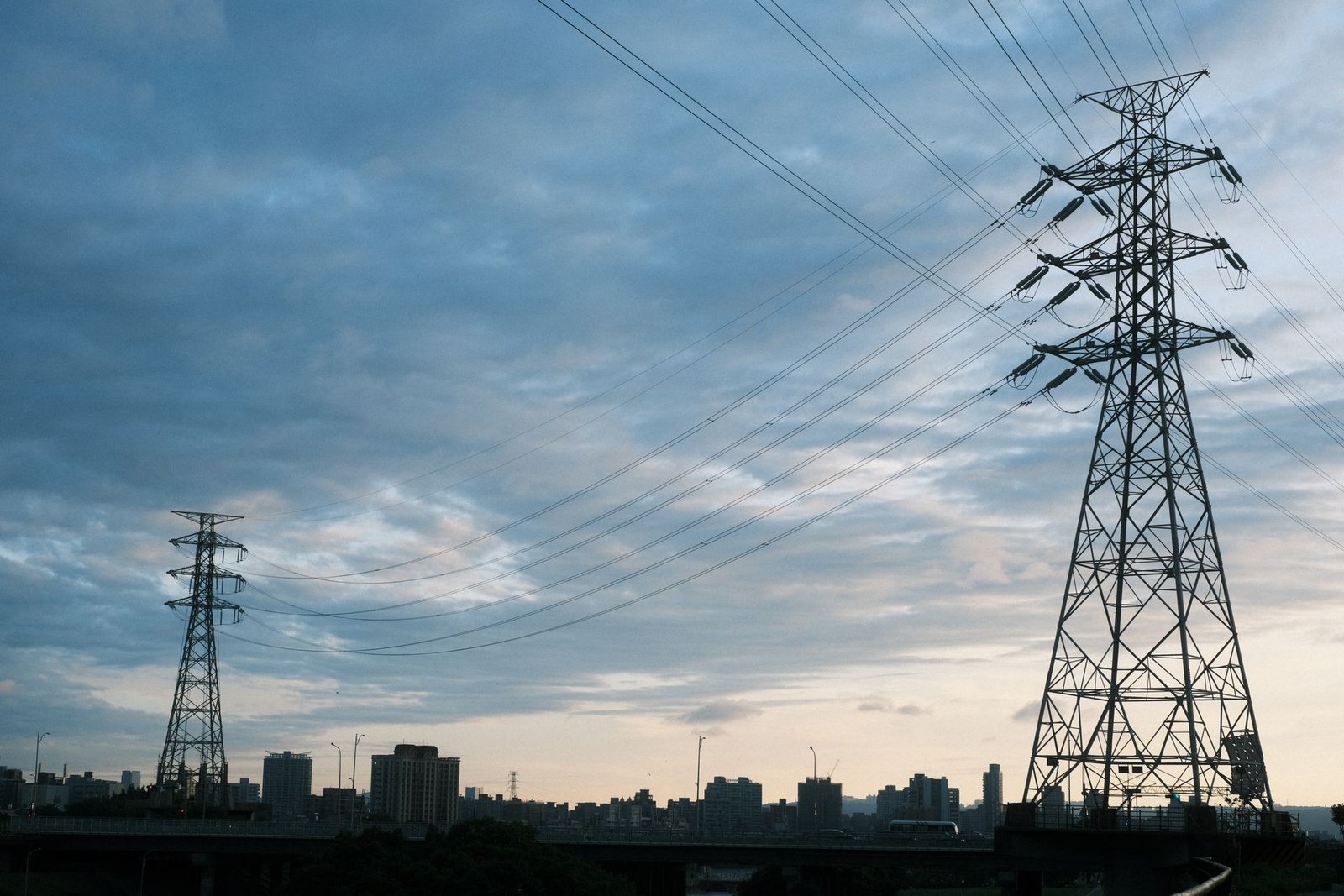 Torres de transmissão de energia elétrica recortadas contra o céu no fim da tarde, com a silhueta de uma cidade ao fundo