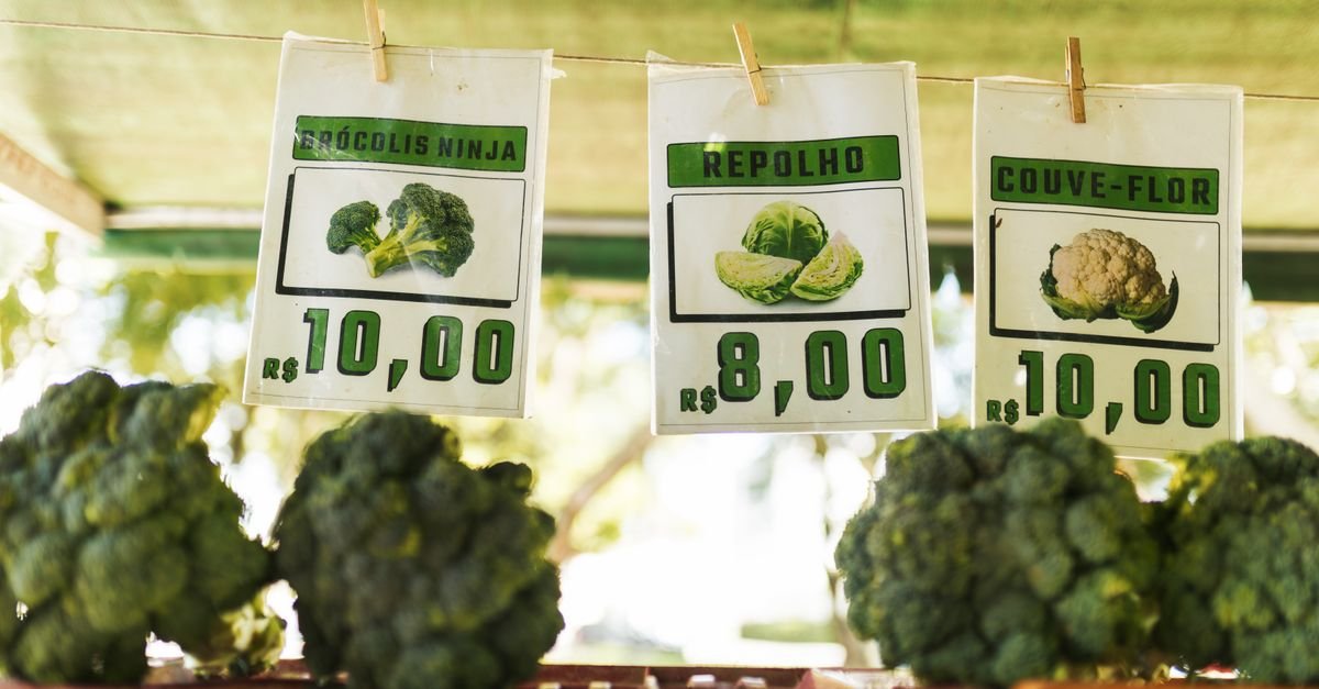 Placas de preco escritas a mao em uma banca de feira, com valores em reais para brocolis, repolho e couve-flor