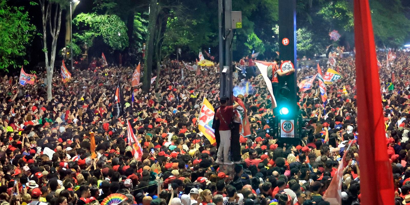 Multidão com bandeiras e camisas vermelhas comemora a reeleição de Lula em rua arborizada, à noite