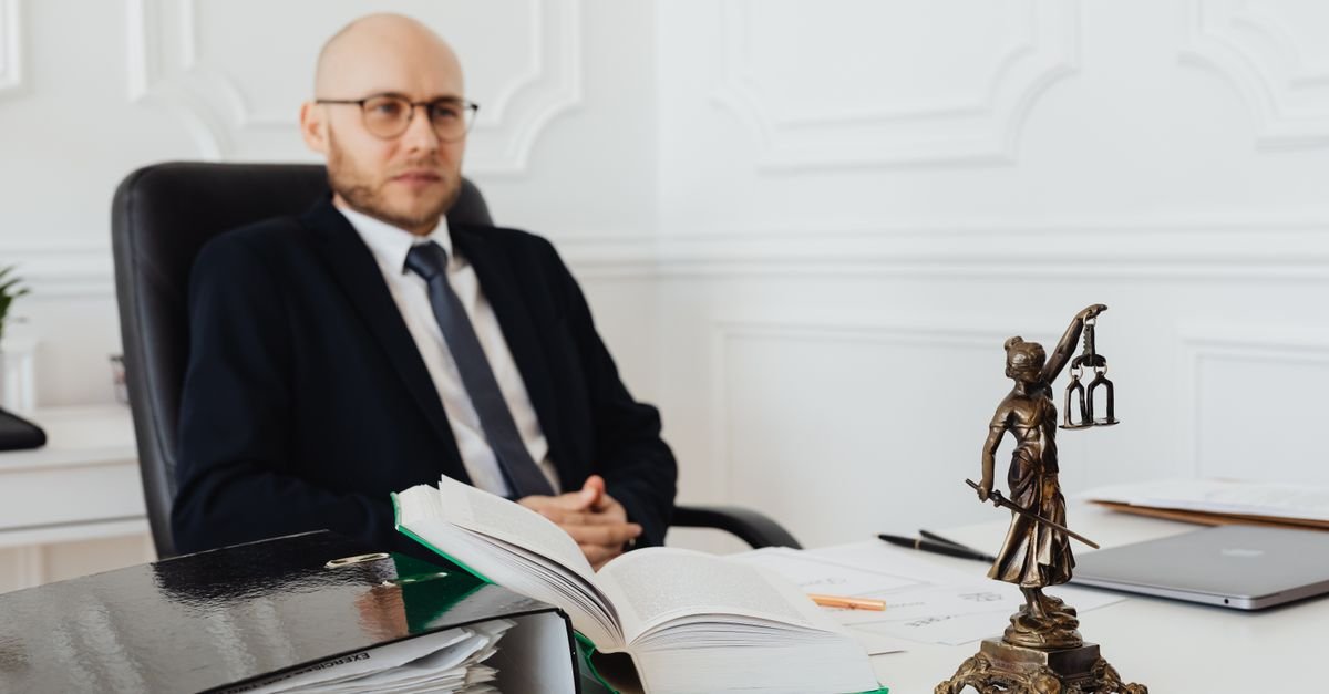 Estátua da Justiça sobre mesa com livro de leis e pastas, advogado desfocado ao fundo