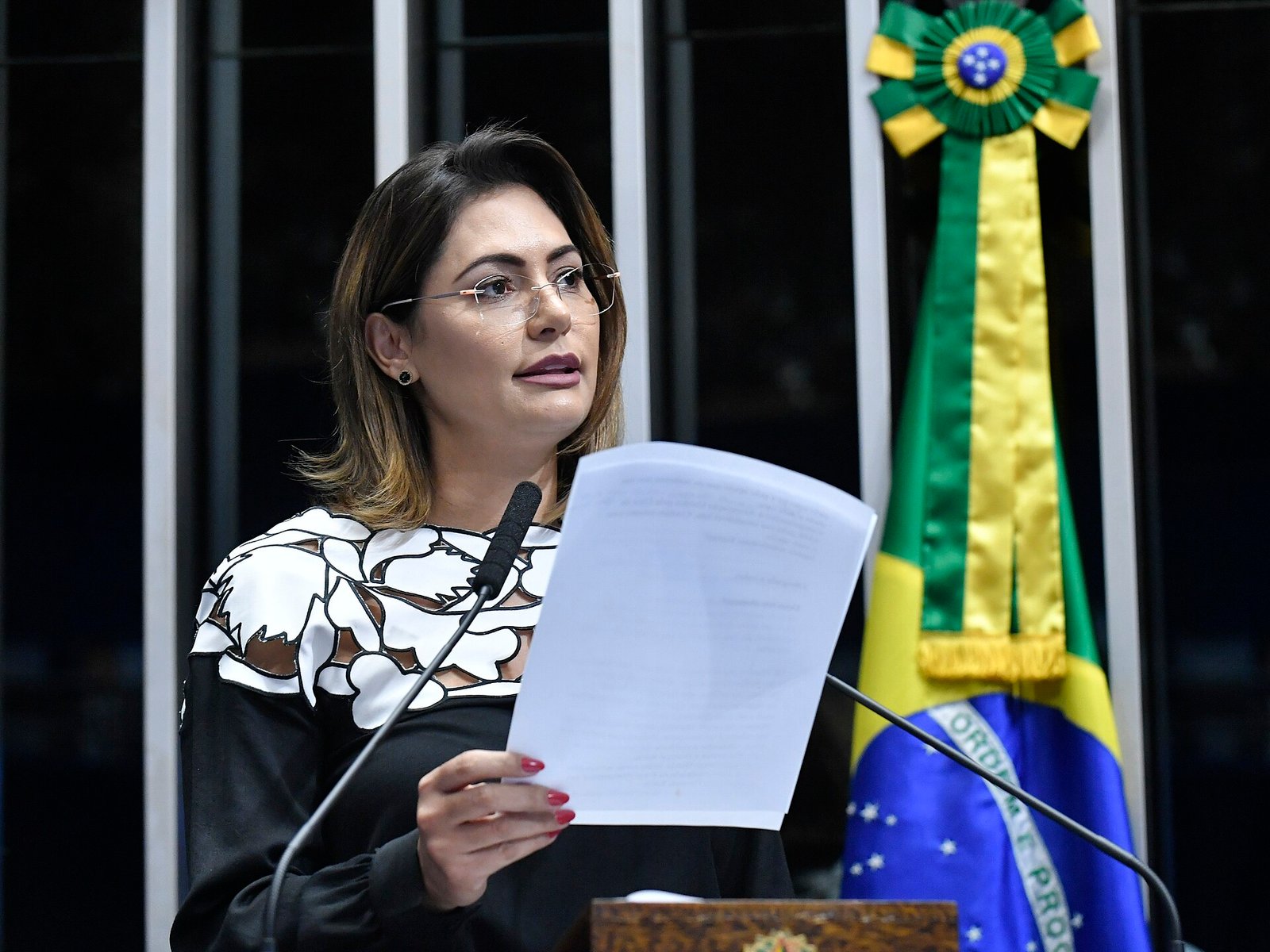 Michelle Bolsonaro na tribuna do plenário do Senado, com microfone e bandeira do Brasil ao fundo