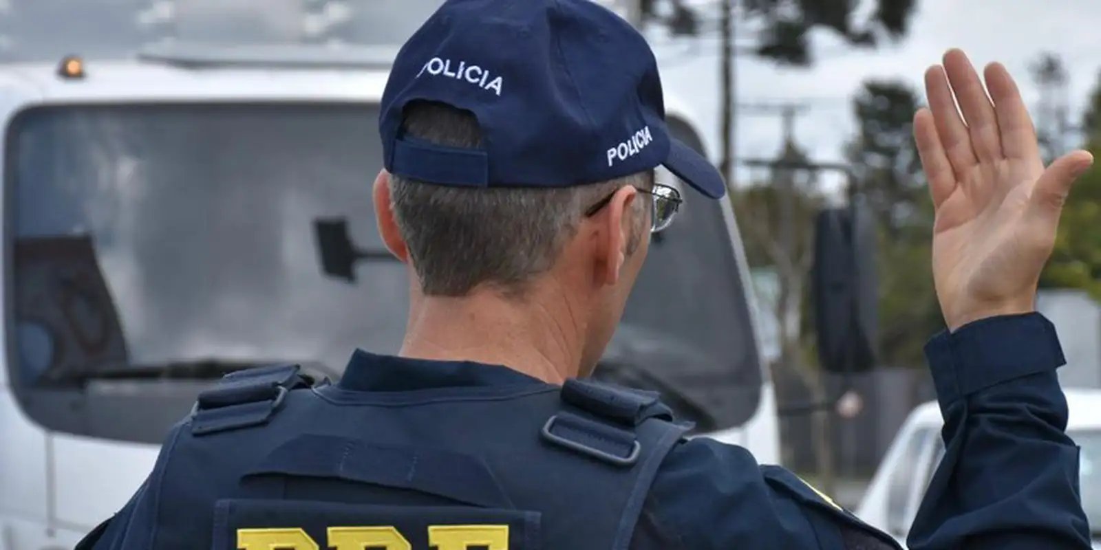 Policial rodoviário federal, de costas, faz sinal de parada com a mão em abordagem na estrada; foto de arquivo