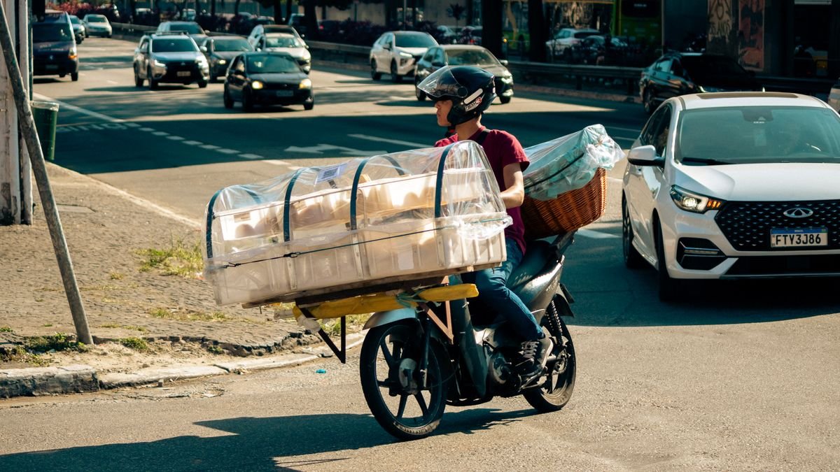 Motociclista transporta caixas plásticas cobertas por lona no trânsito de uma avenida brasileira
