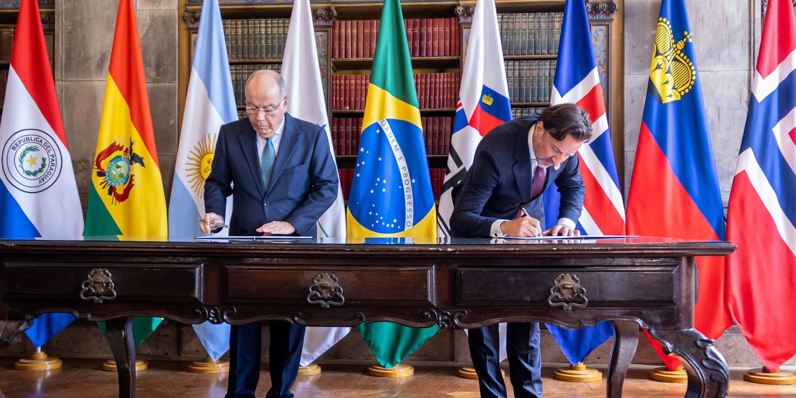 Dois signatários assinam documentos diante das bandeiras dos países do Mercosul e da EFTA, em cerimônia do acordo (foto de arquivo)