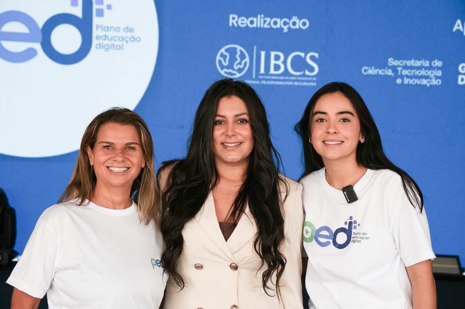Três mulheres sorrindo diante de painel azul com a marca do PED e os logotipos do IBCS e da Secretaria de Ciência, Tecnologia e Inovação