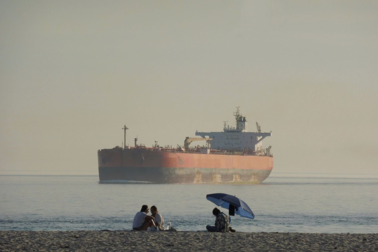 Petroleiro navegando em mar calmo ao largo de uma praia, com neblina ao fundo