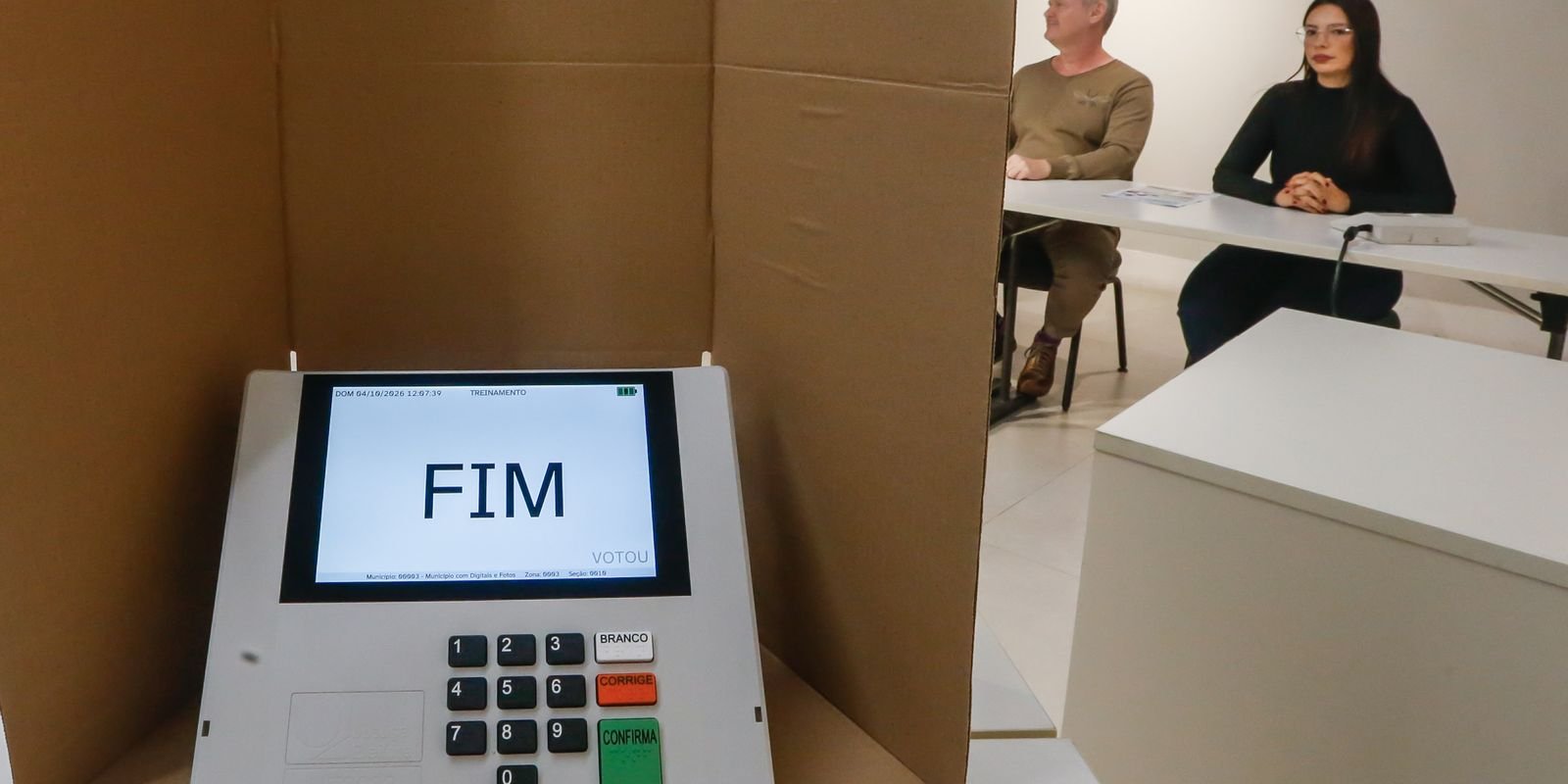 Urna eletrônica em modo de treinamento ao lado de cabine de votação de papelão, em foto de arquivo