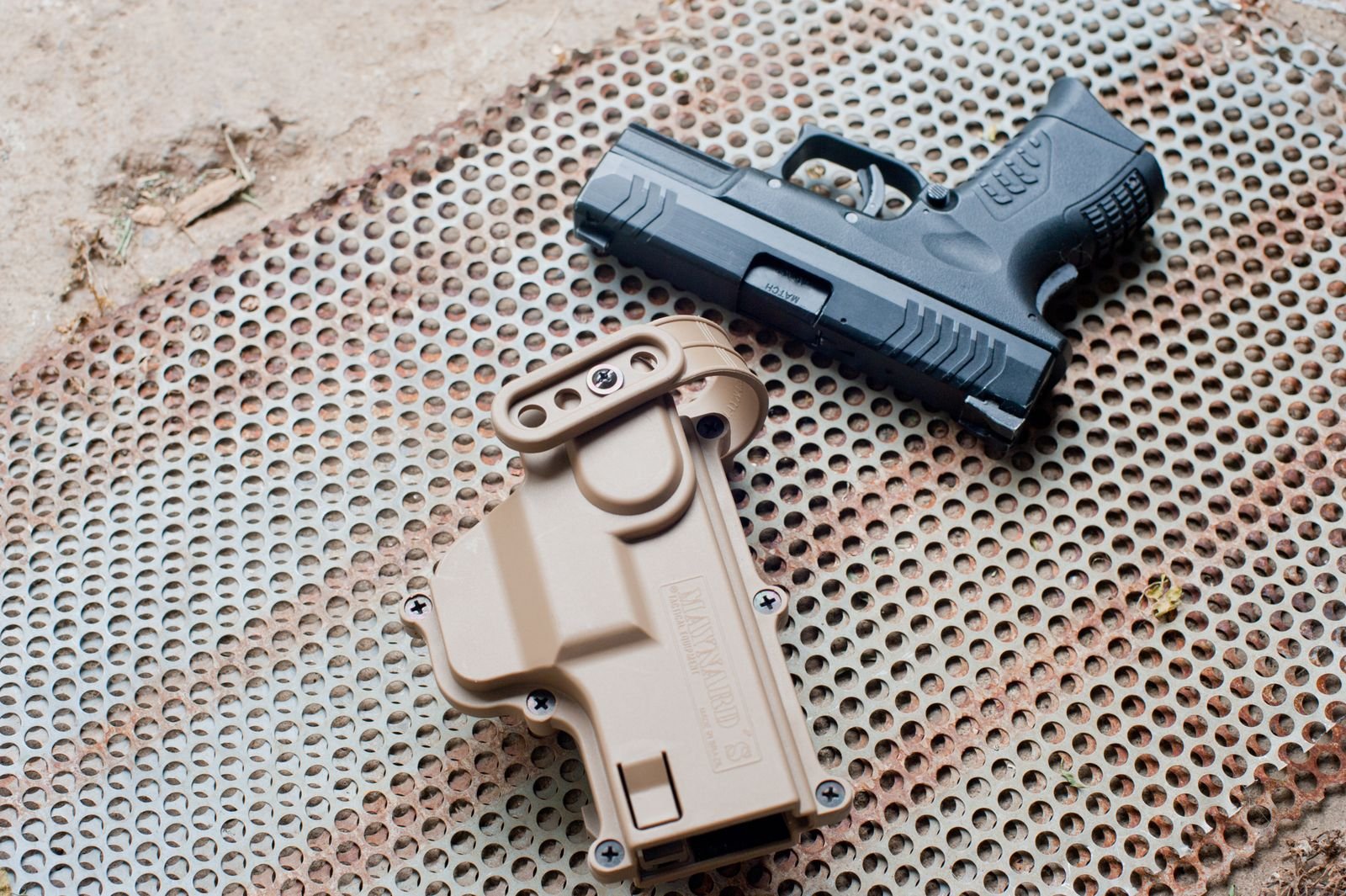 Pistola preta ao lado de um coldre bege sobre superfície metálica perfurada