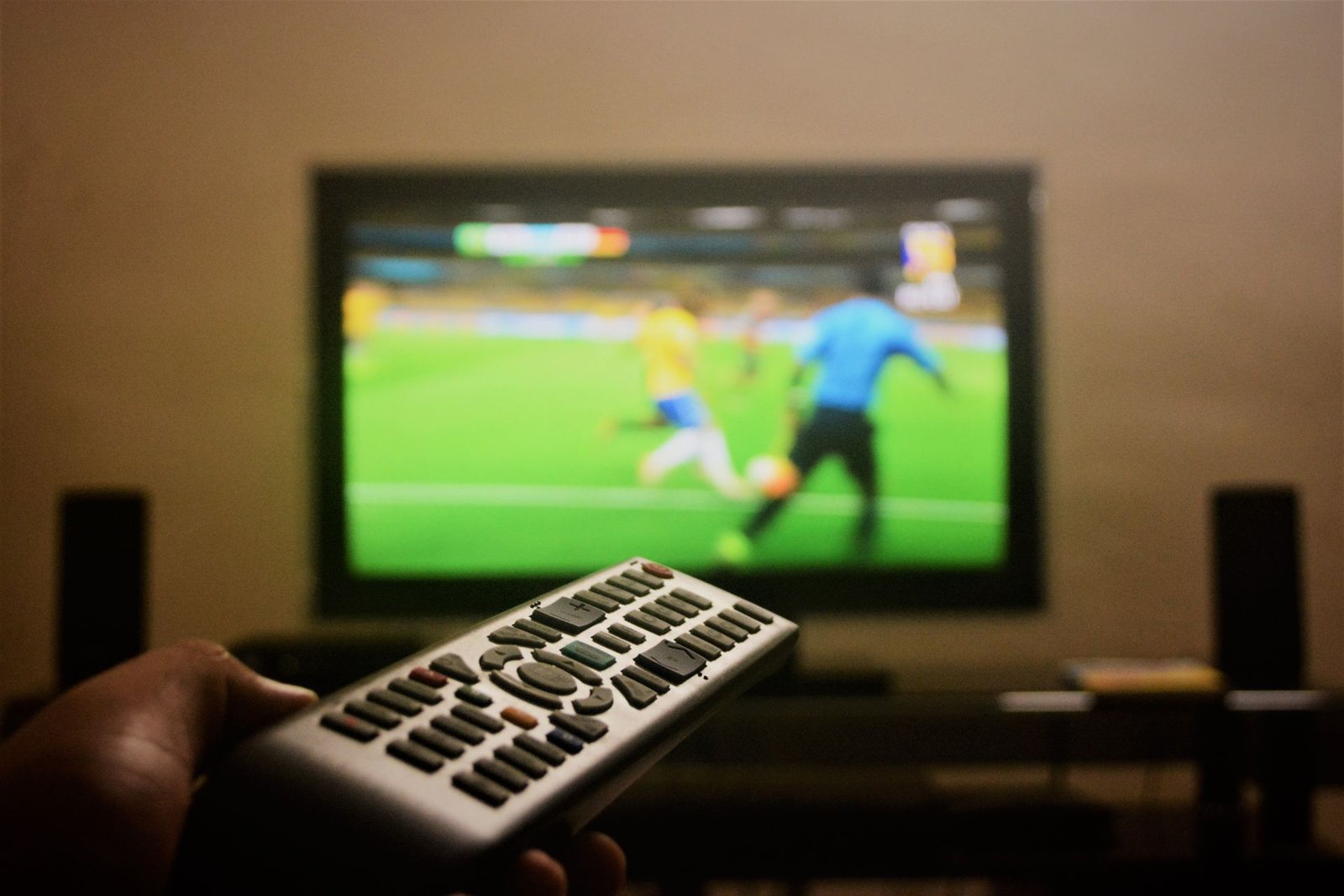 Mão segura um controle remoto em frente a uma televisão que exibe uma partida de futebol