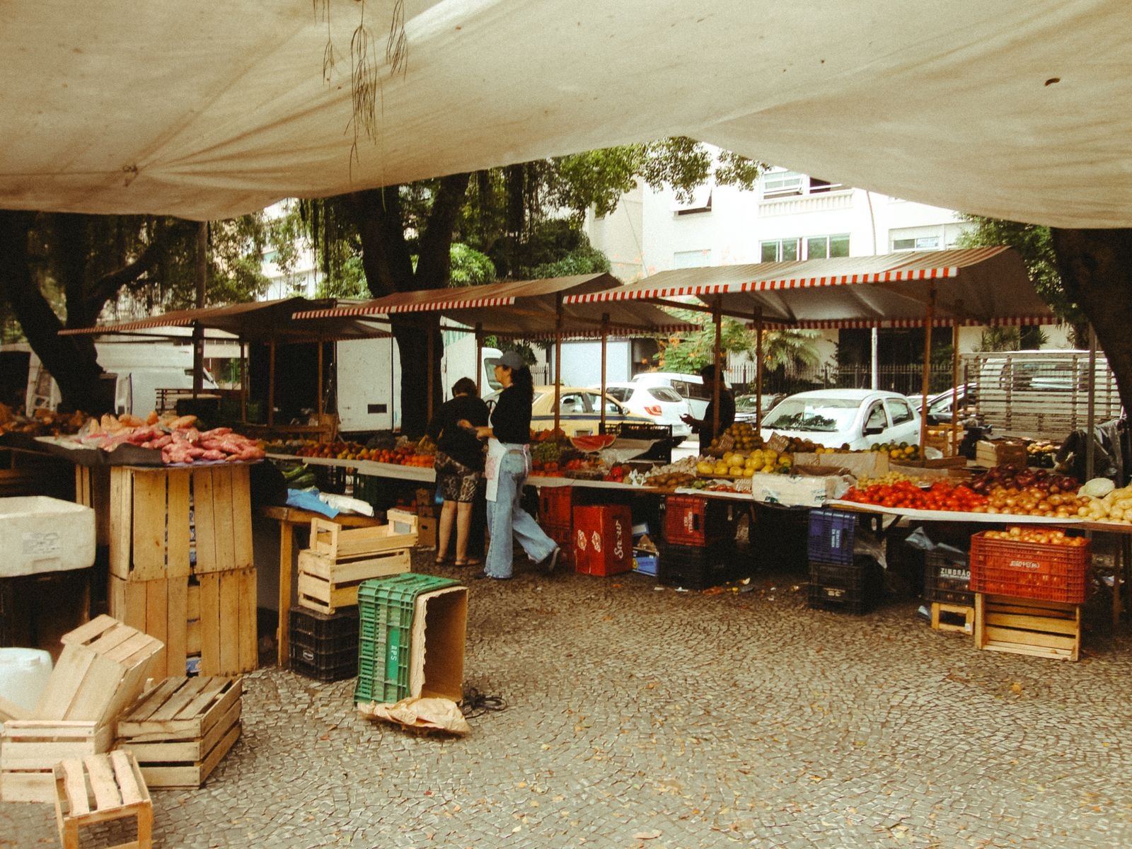 Feira livre com barracas de frutas e verduras, vans estacionadas ao fundo e feirantes atendendo, no Rio de Janeiro