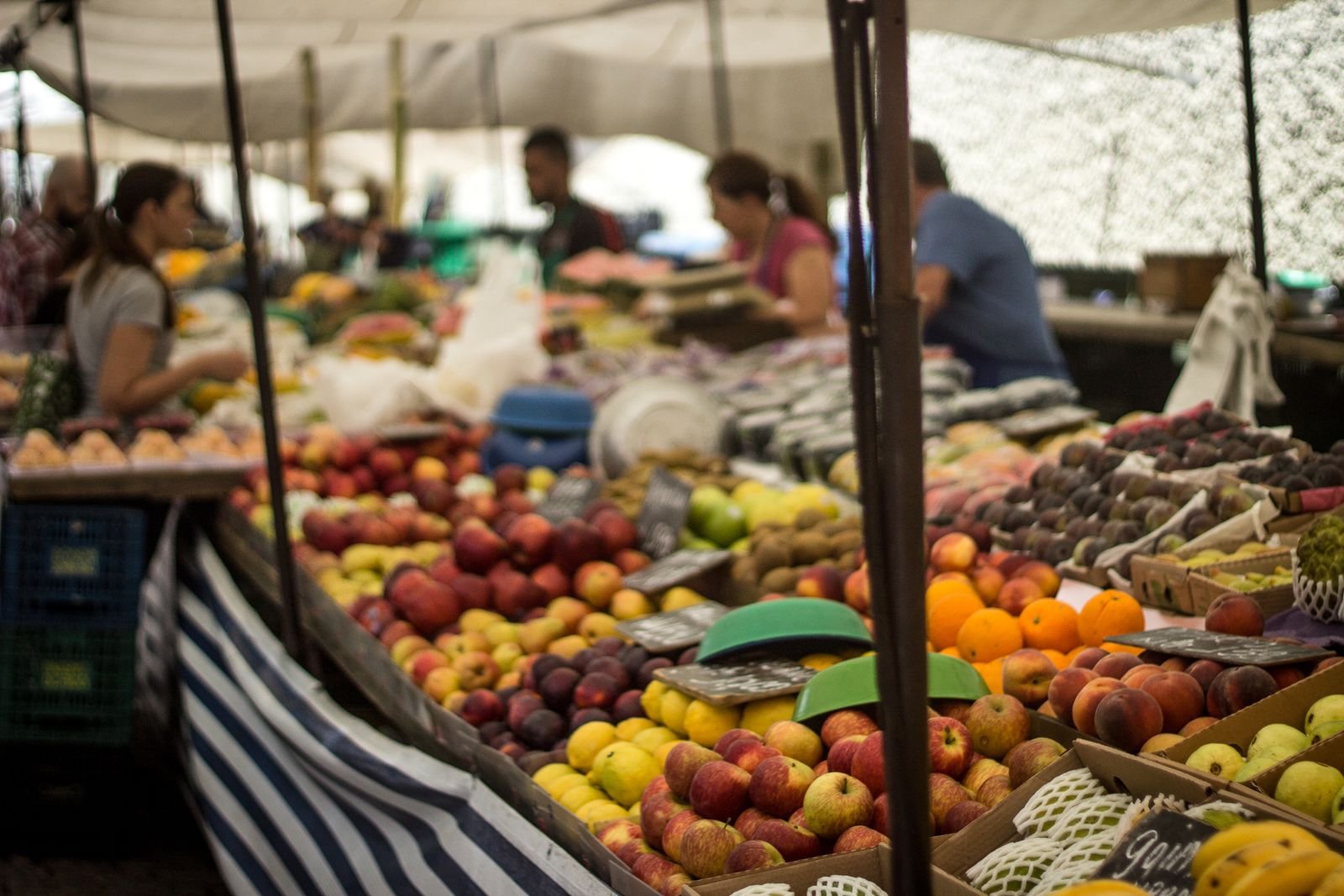 Banca de frutas em feira livre, com maçãs, laranjas e limões expostos e vendedores ao fundo