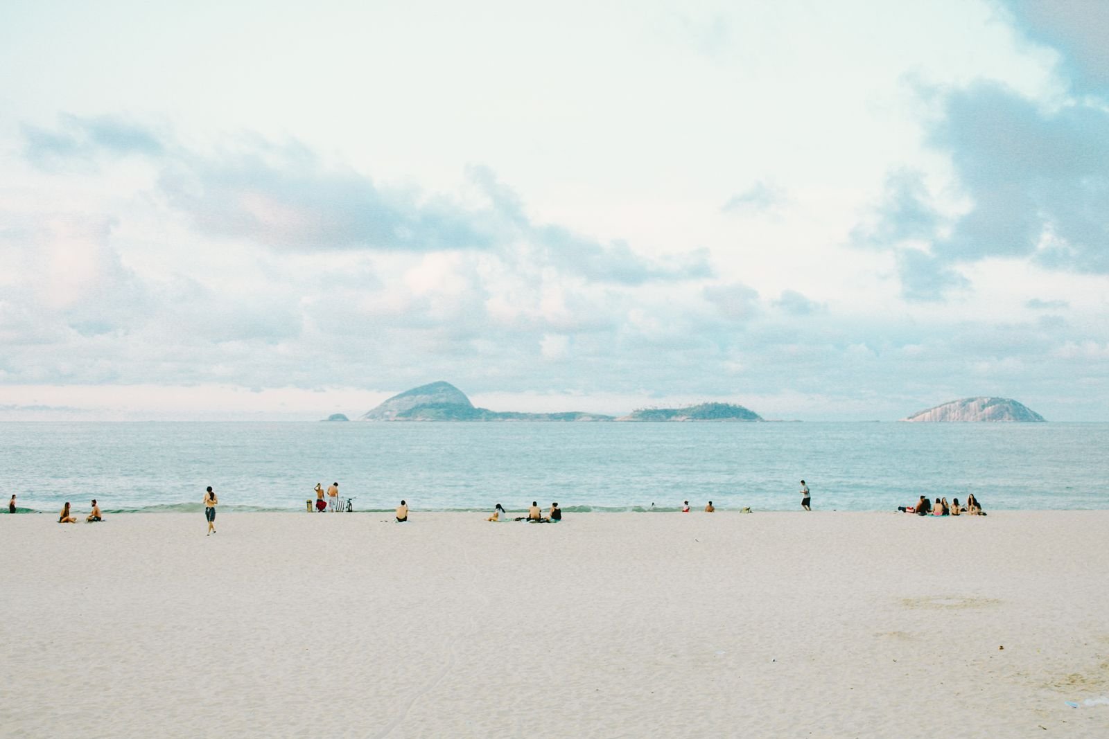 Pessoas sentadas na areia e na beira do mar numa praia do Rio de Janeiro, com as ilhas Cagarras ao fundo