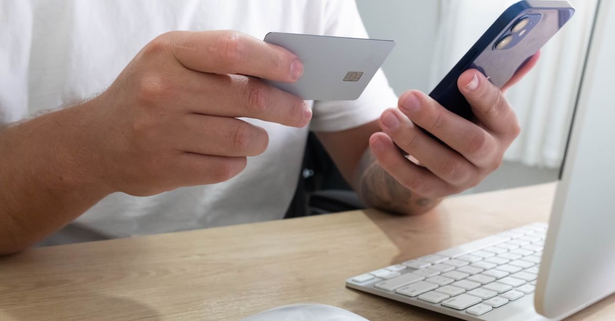 Mãos segurando cartão e celular durante transação online em frente ao computador
