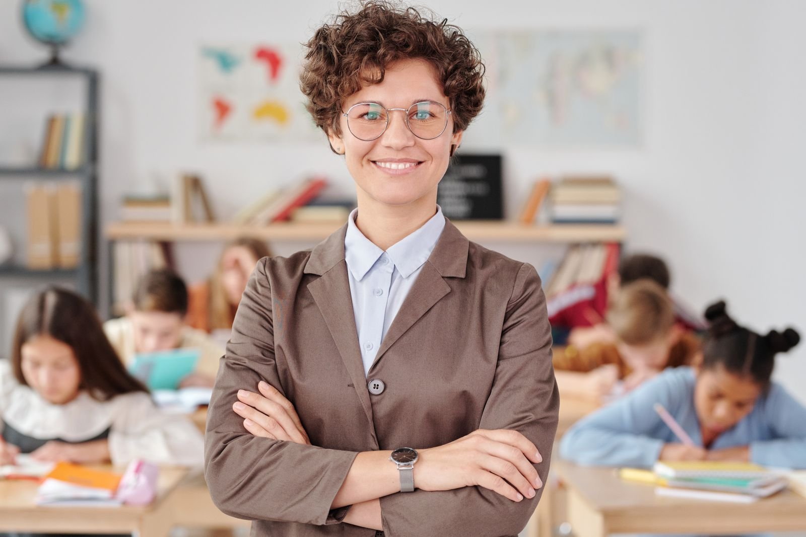 Professora de óculos e blazer marrom sorri de braços cruzados em sala de aula, com alunos ao fundo (foto ilustrativa)